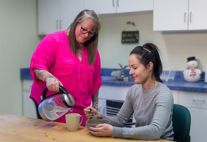 a woman pouring water for another woman in a kitchen.