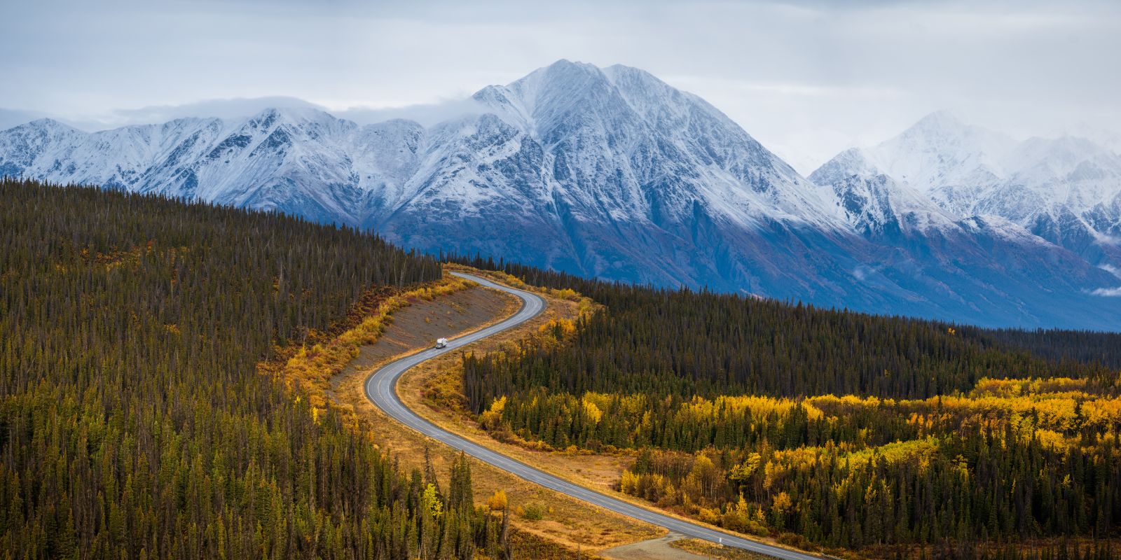 An vehicle towing a trailer drives eastward between Destruction Bay and Haines Junction along the Alaska Highway. Fresh snow blankets Mount Wallace (the high point) and the Kluane Ranges in Kluane National Park and Reserve.