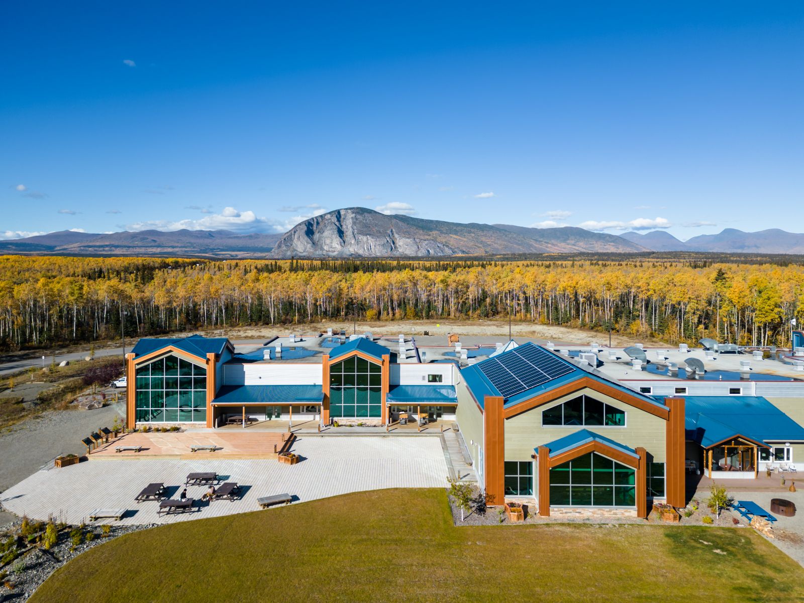 An aerial view of the Da Kų Culture Centre / Haines Junction Visitor Information Centre. The yellow colours of fall are in full force under a clear, blue sky. The prominent bluff of Paint Mountain protrudes above the distant horizon.