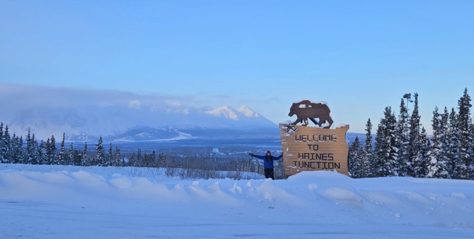 A travelling physiotherapist poses next to the Welcome to Haines Junction sign in Haines Junction, Yukon