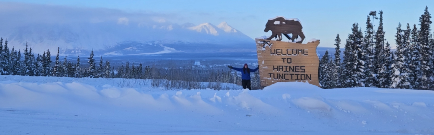 A travelling physiotherapist poses next to the Welcome to Haines Junction sign in Haines Junction, Yukon